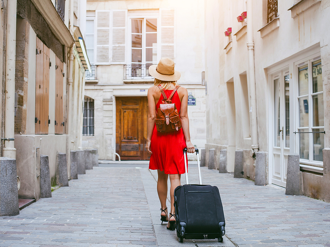 Woman rolling suitcase through narrow street, seen from behind
