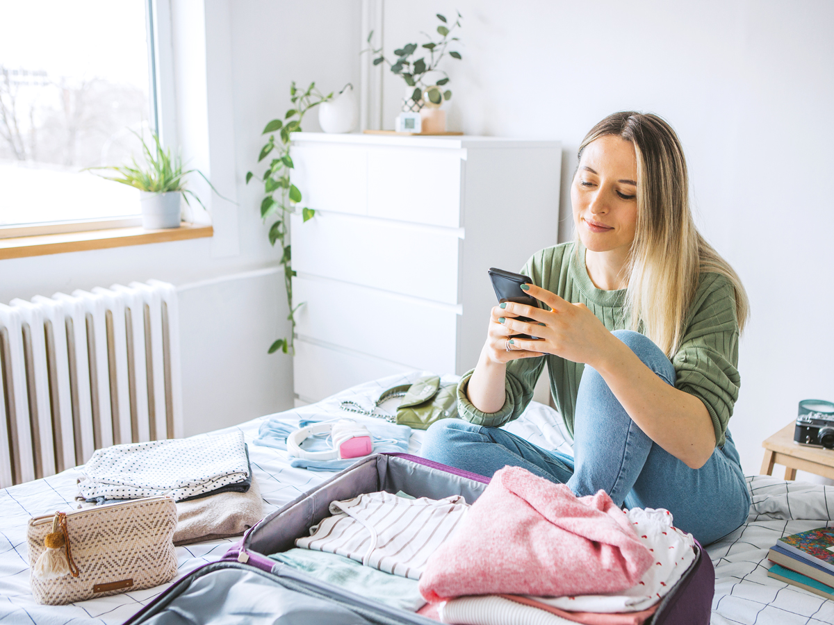 Traveler looking at cellphone while sitting on bed and packing suitcase