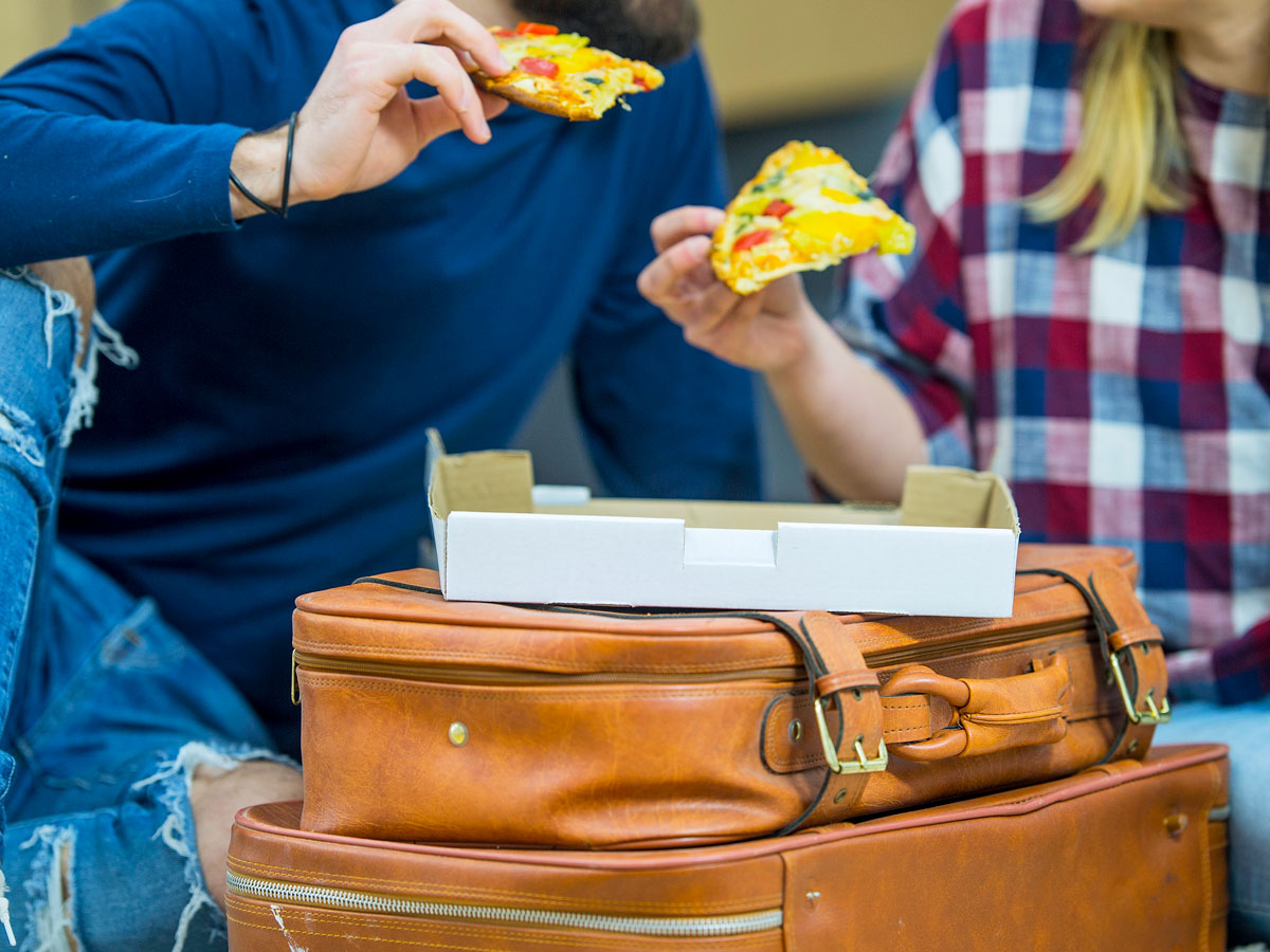 Two people eating pizza from box on top of luggage