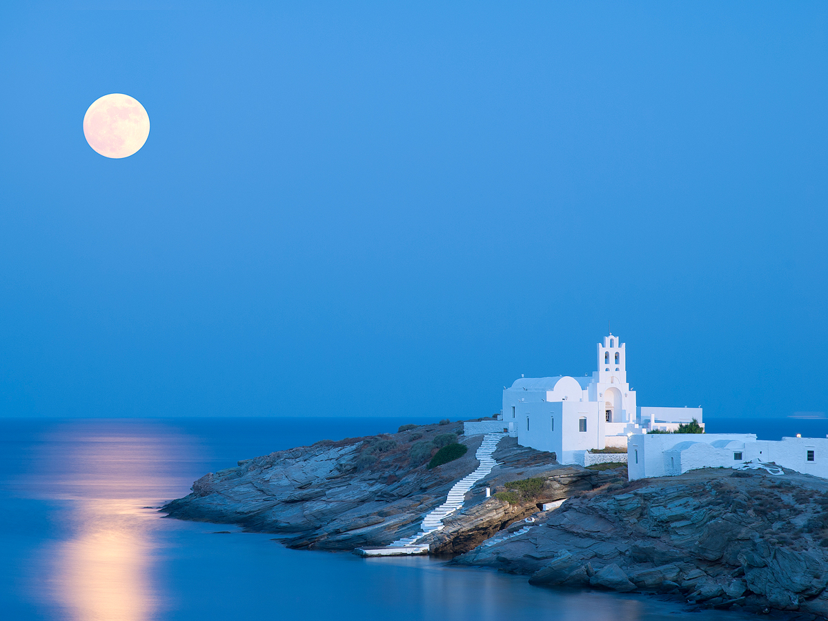 White church on rocky peninsula with moon visible on Sifnos, Greece